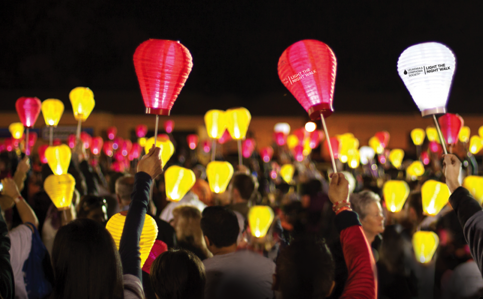 A crowd at nighttime holding up illuminated paper lanterns in various colors, with some lanterns displaying logos and text.
