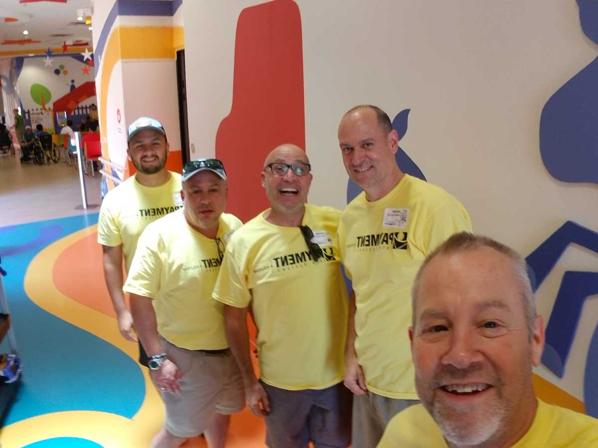 Four smiling men in matching yellow shirts labeled "teammates" take a selfie in a colorful room with abstract shapes on the walls.