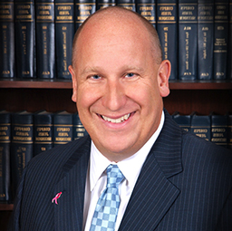 Bradley Rollins, a smiling bald man wearing a dark suit, light blue checked tie, and a pink lapel pin, posing in front of a bookshelf filled with law books.