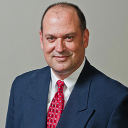 A professional headshot of a middle-aged man with a subtle smile, wearing a dark blue suit, white shirt, and red tie with circular patterns.