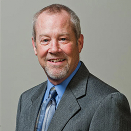 A professional portrait of a smiling older man with a beard, wearing a gray suit and a blue tie against a neutral background.