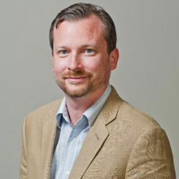 A professional headshot of a smiling man with a beard, wearing a beige blazer over a blue shirt.