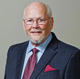 Elderly man with a white beard, wearing a smart navy blazer, red patterned tie, and a light blue shirt, standing against a gray background. He has a gentle smile. This