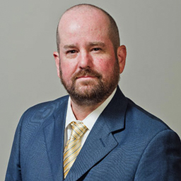 A professional headshot of Miguel A. Chapa, a bald man with a beard, wearing a blue suit, white shirt, and yellow striped tie. He is looking directly at the camera with a
