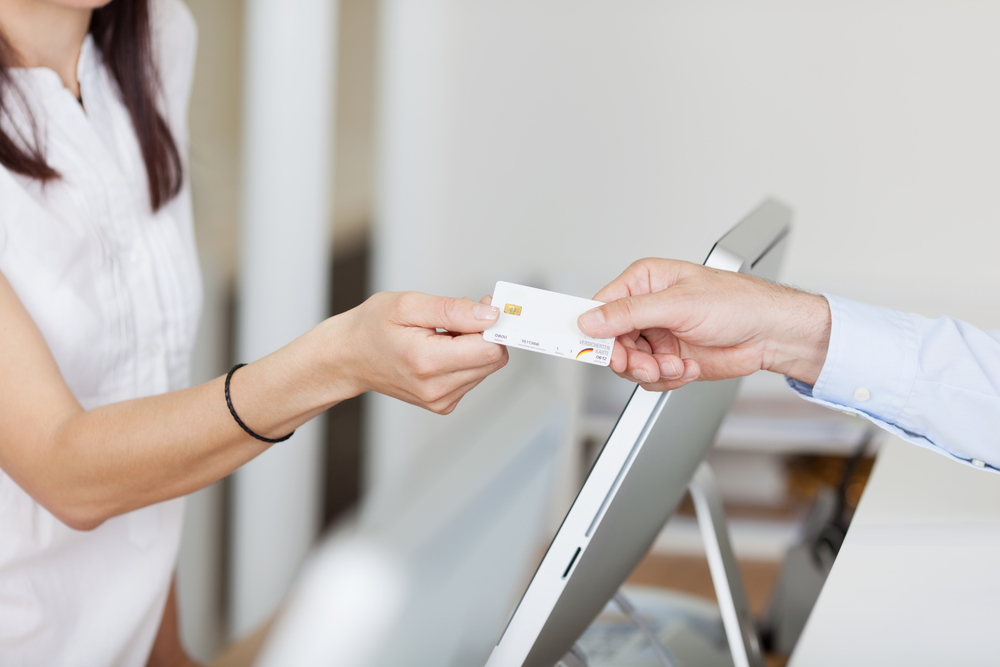 A customer hands a credit card to a receptionist at a modern service desk, representing a payment or check-in process in a clean and well-lit office environment.